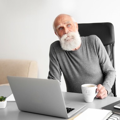 Elderly man sat by laptop with a cup of tea.