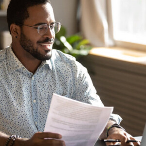 A man sitting at a desk in his home office. He's holding a report document and looking at his laptop.