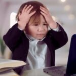 Worried toddler in office with laptop, holds head in hands