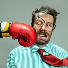 Business man grimacing as boss wearing boxing glove punches him in the face